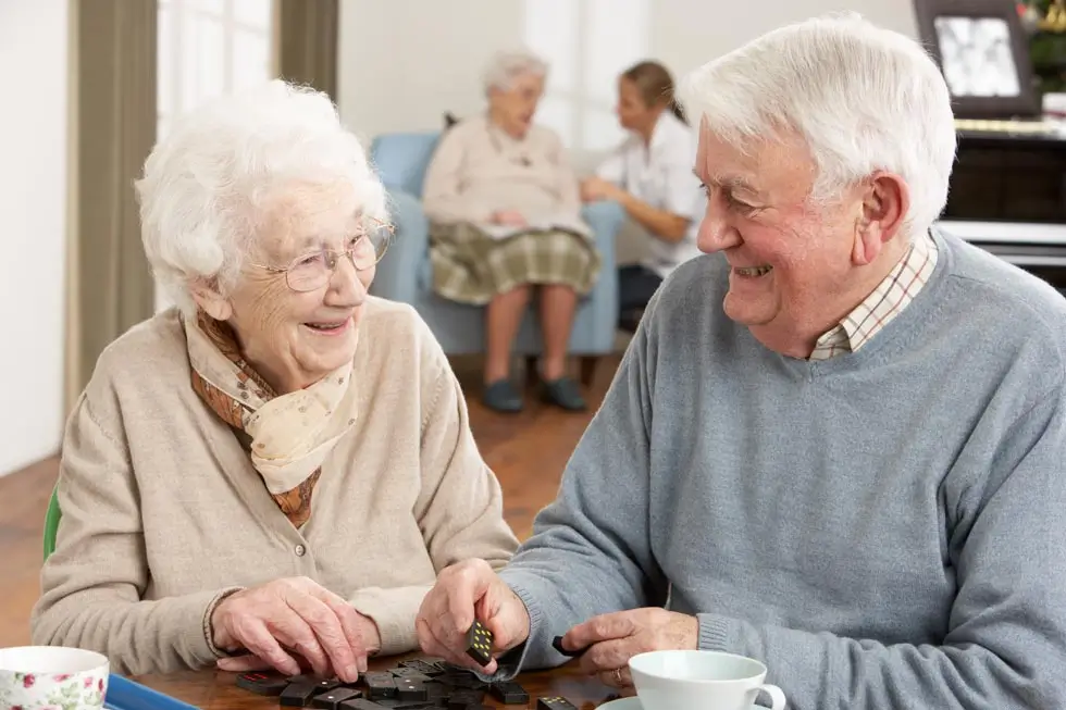 Zwei ältere Personen sitzen an einem Tisch und spielen Domino, im Hintergrund sitzen zwei weitere Personen auf einem Sofa.