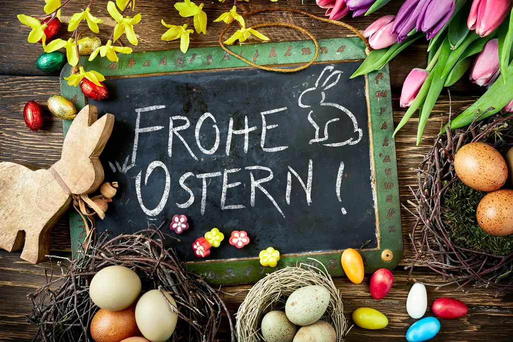 Tafel mit der Aufschrift 'FROHE OSTERN' und gezeichnetem Hasen umgeben von Osterdeko wie Holzhasen, bunten Eiern, Tulpen und Frühlingszweigen