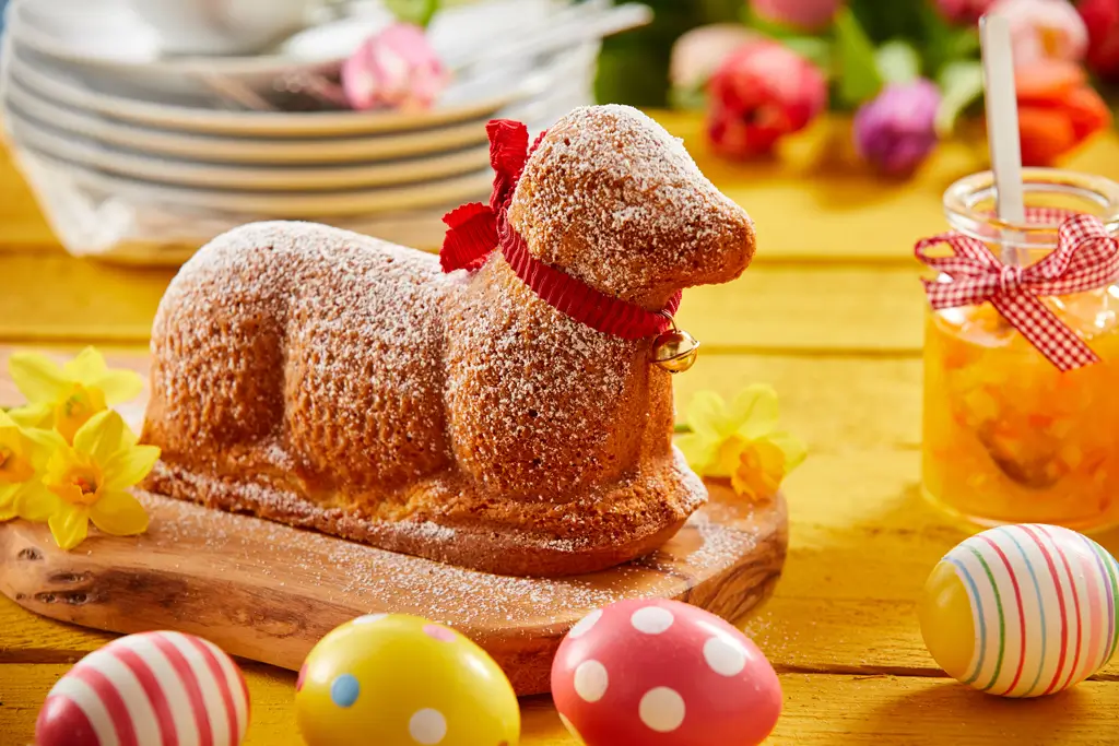 Osterlamm aus Kuchen mit Puderzucker bestäubt und rotem Band mit Glöckchen auf Holzbrett umgeben von bunten Ostereiern und Frühlingsblumen