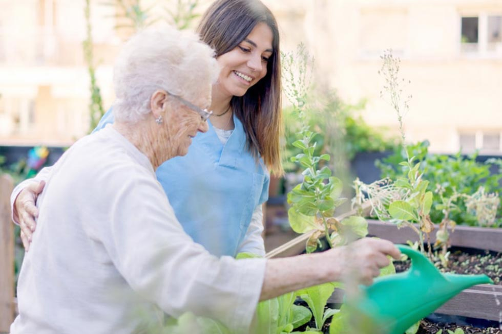 Pflegekraft und Seniorin gießen gemeinsam Pflanzen in einem Hochbeet im Freien